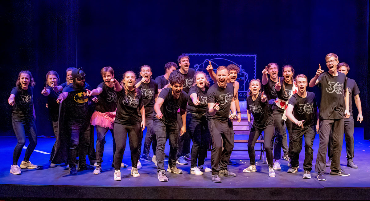 A group of Bethany Lutheran College students are on stage in matching shirts, energetically posing and pointing toward the camera under bright stage lights.