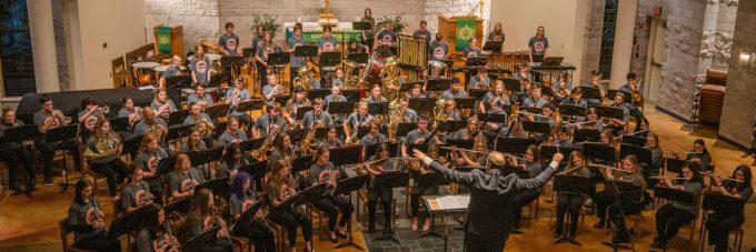 A conductor leads the Bethany Lutheran College concert band as students perform on stage in a brightly lit church or auditorium.