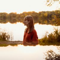 A woman in a red sweater stands beside a Minnesota lake at sunset, with her reflection visible on the shimmering water.