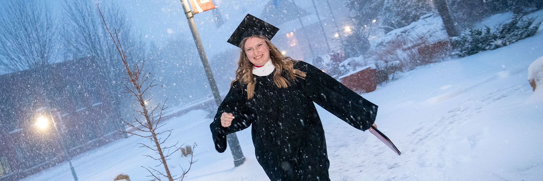 A Bethany Lutheran College student in graduation attire walks outside in falling snow, holding a diploma and smiling.
