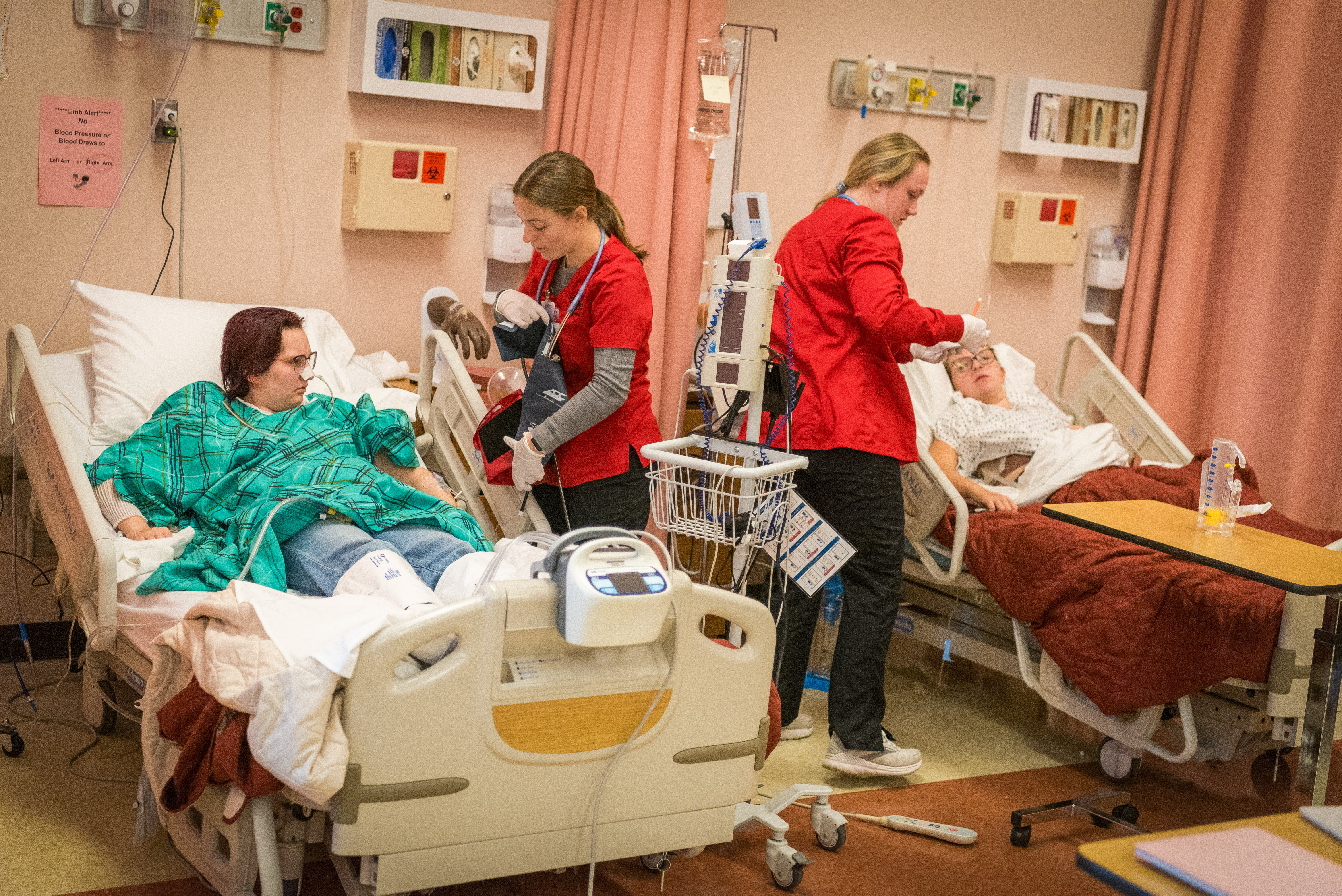 Two nurses in red uniforms care for two patients lying in hospital beds in a medical room with medical equipment. This image could be used to illustrate healthcare training or medical scenarios; if related to Bethany Lutheran College, it may represent students participating in a nursing or allied health program.