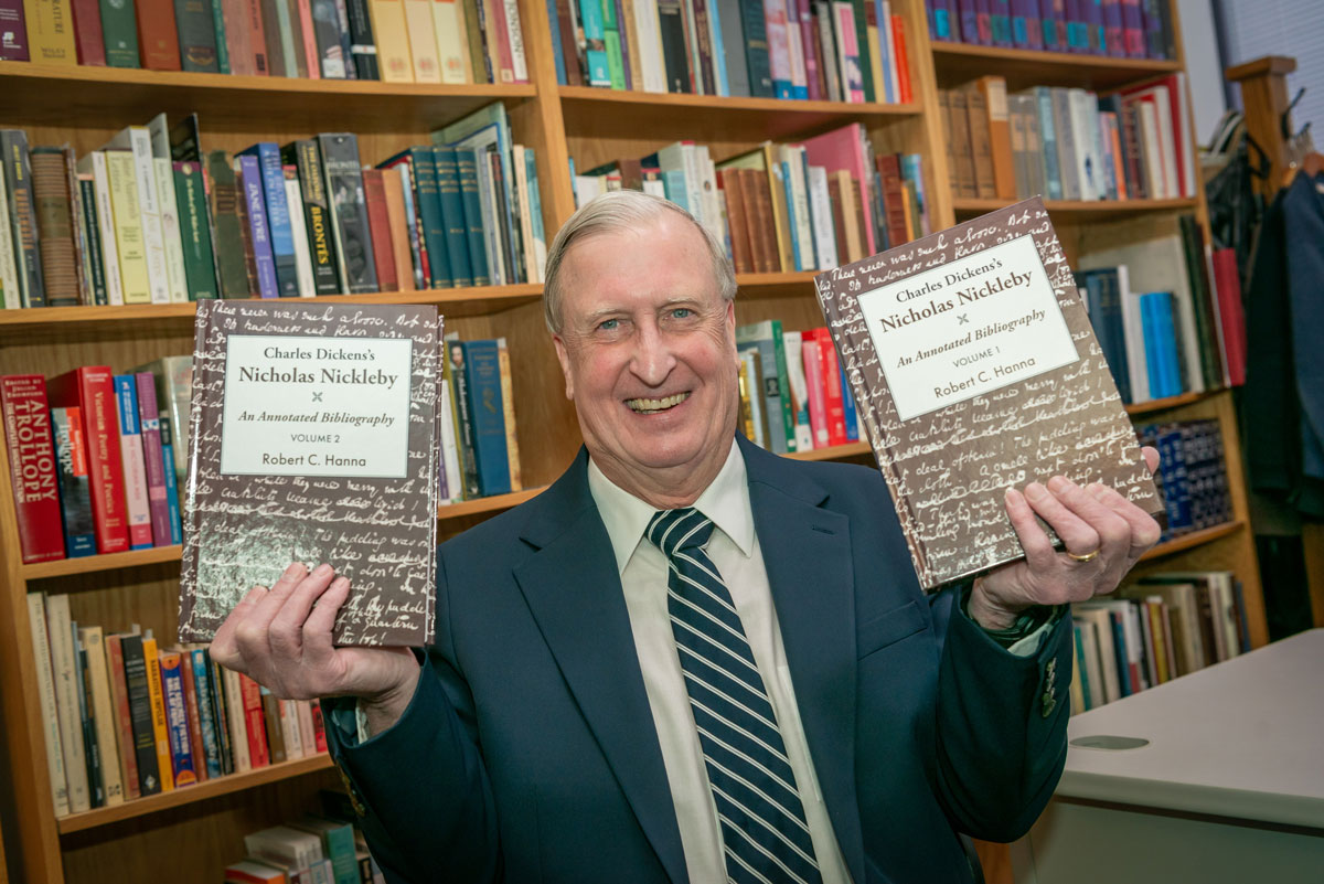 A Bethany Lutheran College faculty member in a suit smiles while holding two large books, standing in front of a bookshelf filled with various titles.