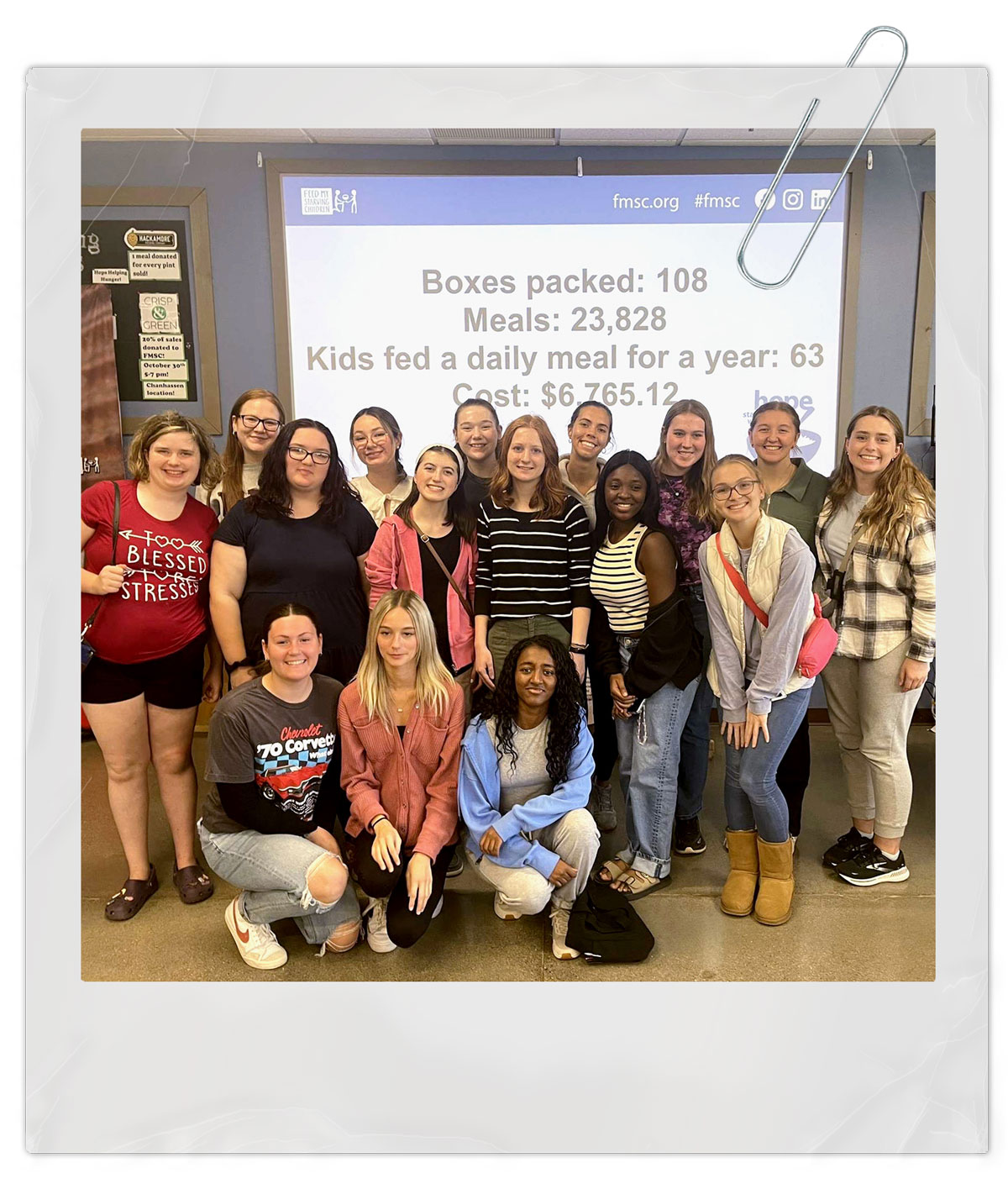 Bethany Lutheran College students and community members pose in front of a screen displaying food packing statistics and cost at an indoor event.