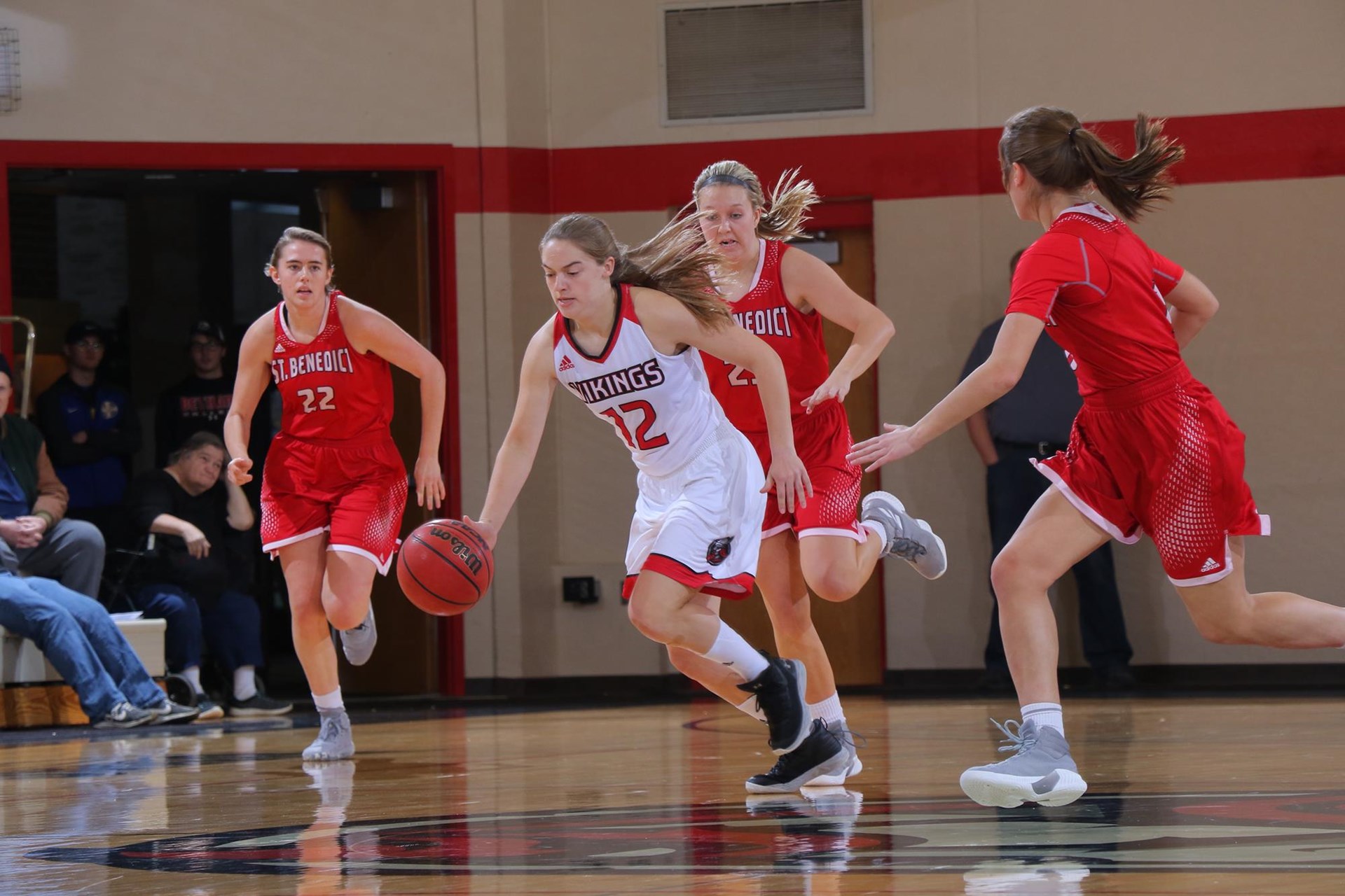 A Bethany Lutheran College player in a white uniform dribbles past three defenders on an indoor basketball court in Mankato.