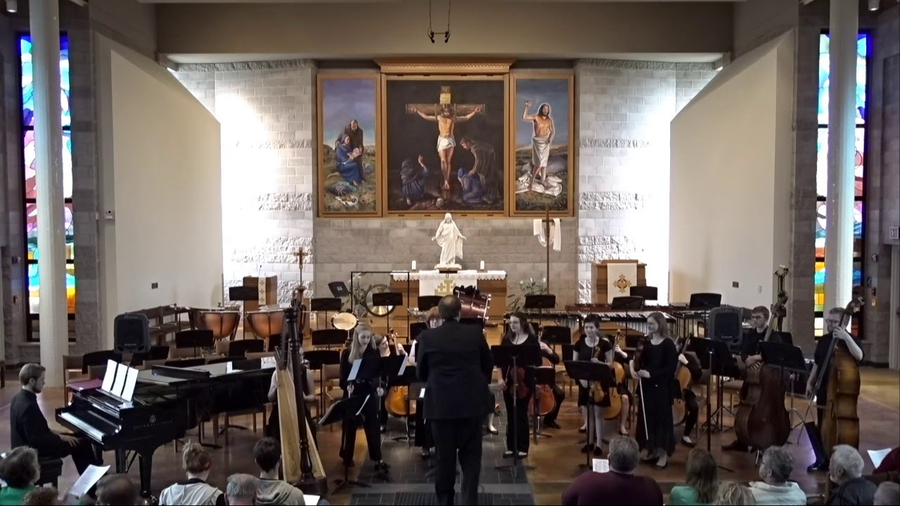 A chamber orchestra from Bethany Lutheran College performs in a church, with stained glass windows visible in the background and students actively engaged in the music.