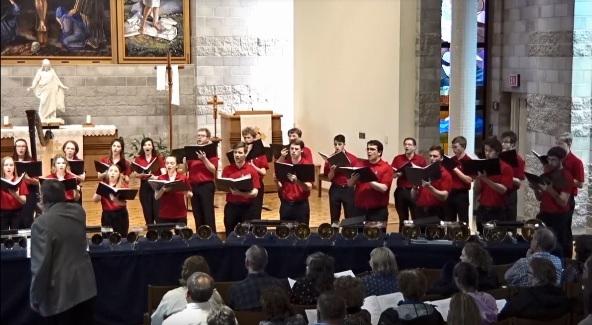 A choir wearing red shirts sings at Bethany Lutheran College in Mankato, with bells arranged in front of them and the audience seated facing the performers.