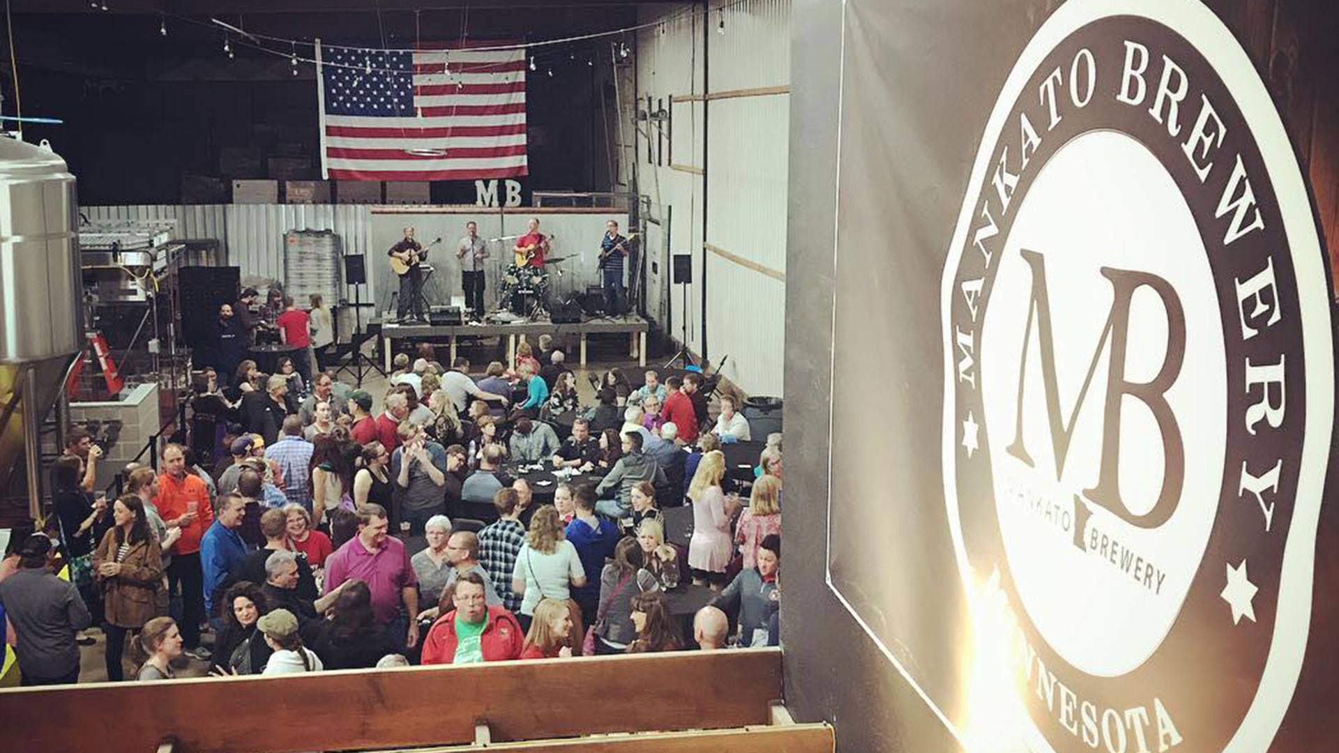 A crowd gathers at Mankato Brewery in Minnesota, watching a band perform on stage with an American flag displayed in the background.