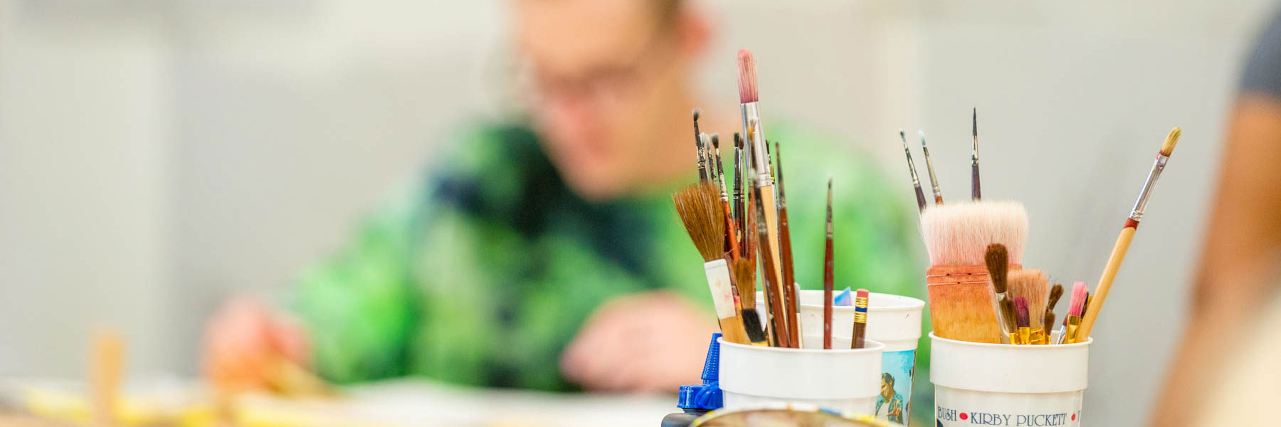 Assorted paintbrushes in cups on a table, with a Mankato student painting in the blurred background.