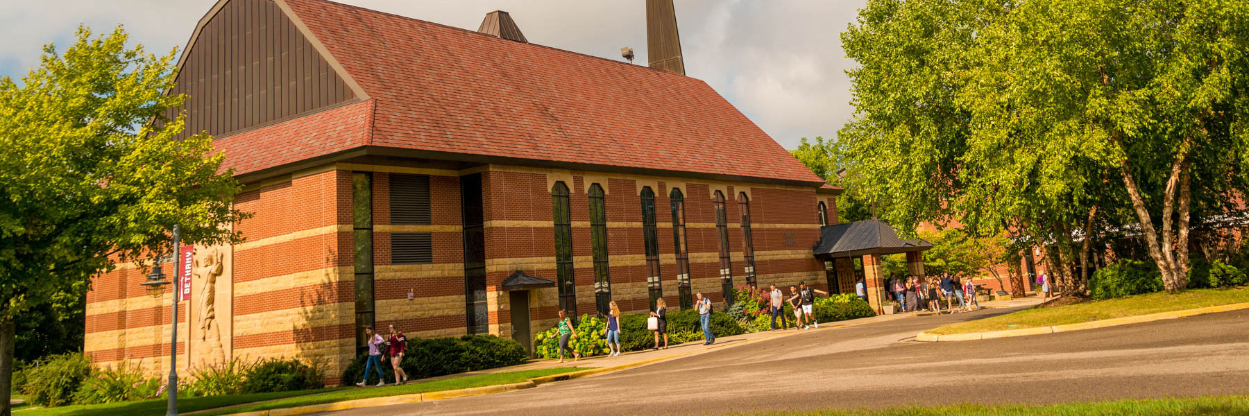 Red-brick building with arched windows at Bethany Lutheran College; students walk outside among green trees on a sunny day.
