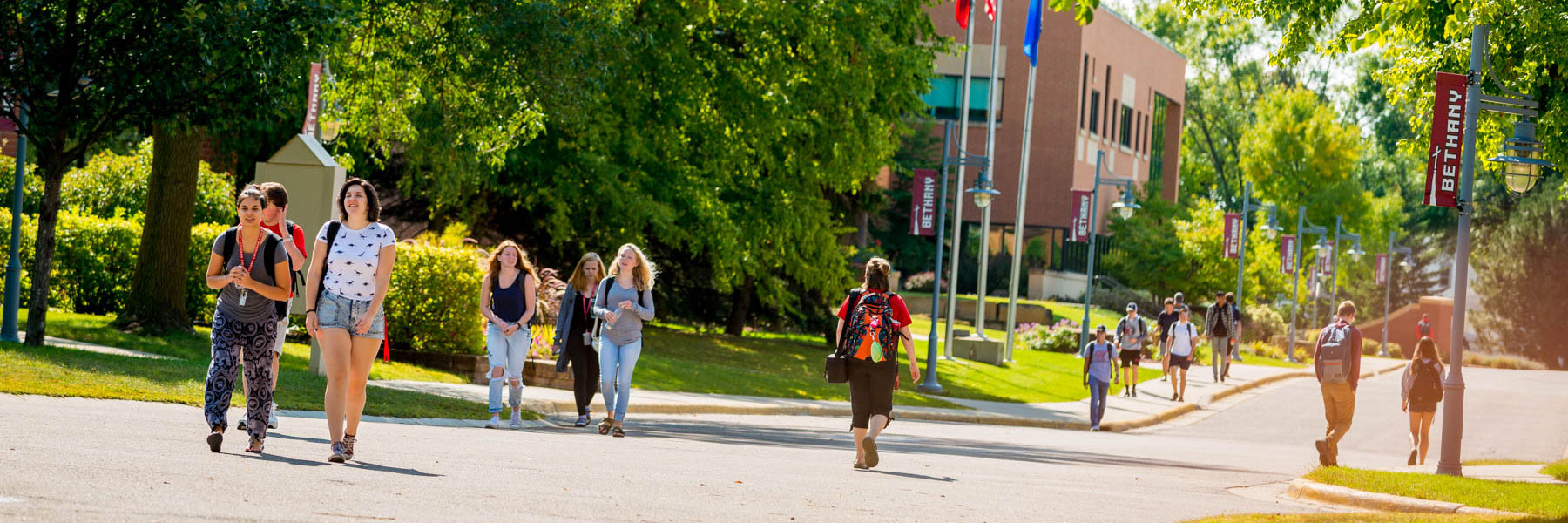 Bethany Lutheran College students stroll along a tree-lined campus pathway adorned with banners on a sunny day in Mankato.