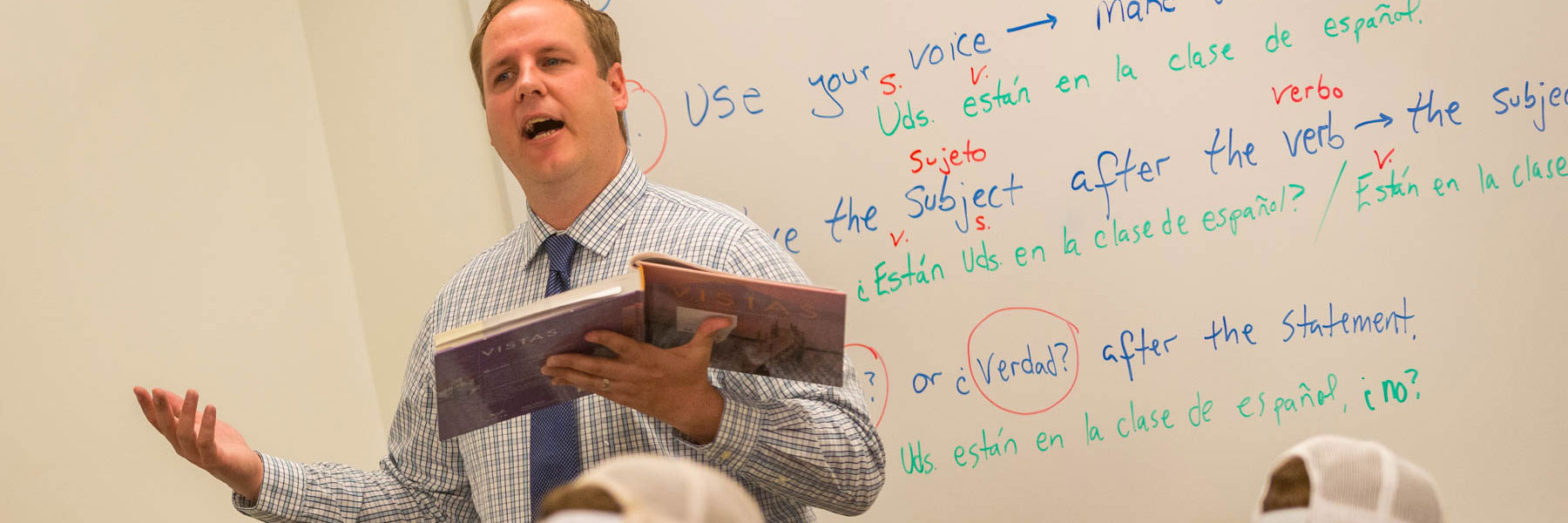 A teacher holding a book speaks in front of a whiteboard displaying Spanish grammar notes, engaging students.