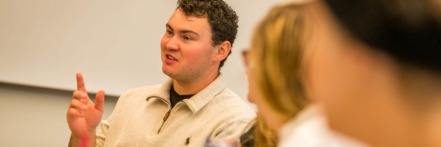 A man with curly hair wearing a beige sweater gestures while speaking to members of the campus community at Bethany Lutheran College.