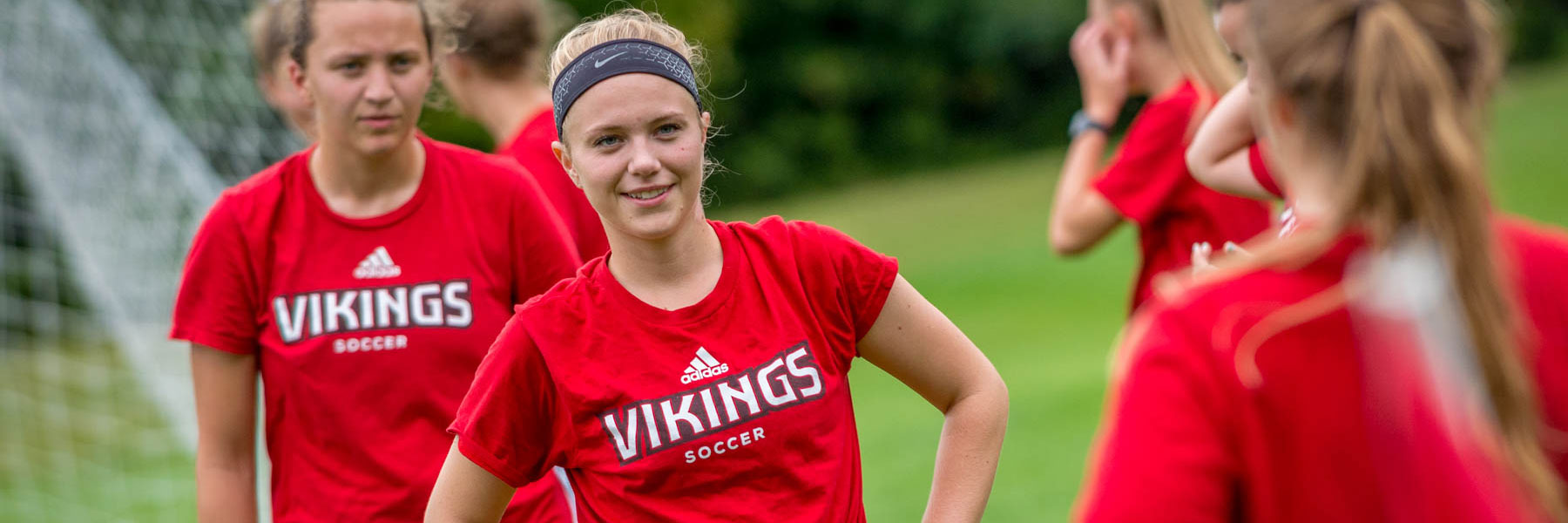 Female soccer players wearing red Vikings Soccer shirts stand on a field in Minnesota, smiling and conversing during practice.