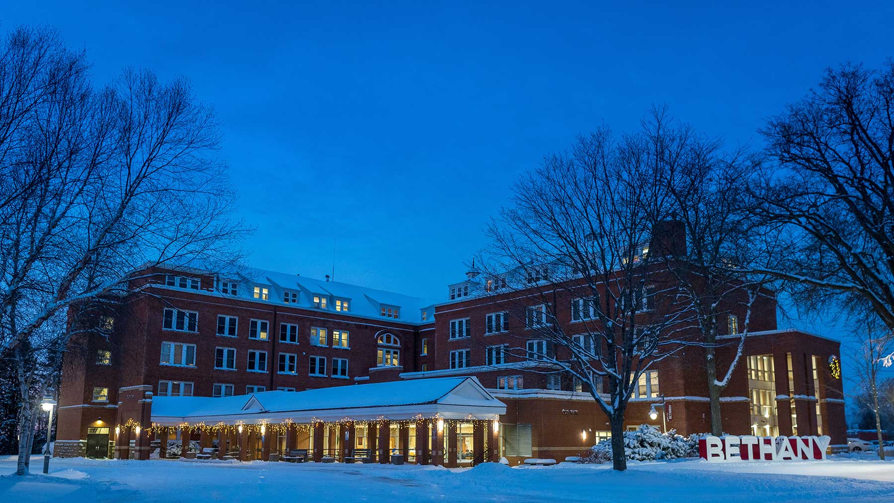 A large brick building at dusk is surrounded by snow and trees, with lit windows. A Bethany Lutheran College sign welcomes the campus community.