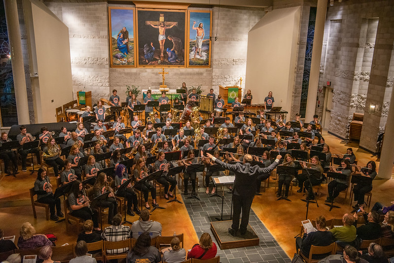 A large band performs in a church in Mankato, led by a conductor, with campus community members seated in the audience.