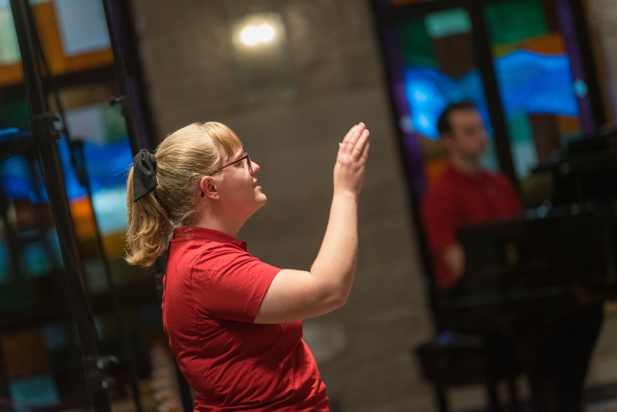 A woman in a red shirt conducts alongside a pianist, encouraging student engagement at Bethany Lutheran College.