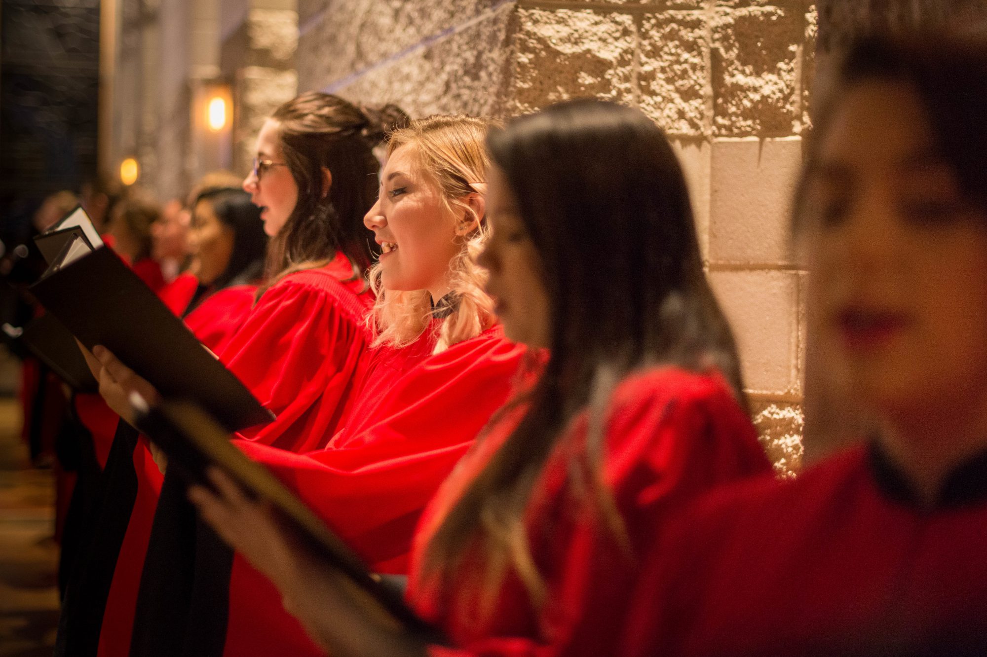 A choir of women in red robes sings together while holding black folders, standing in front of the stone wall at Bethany Lutheran College.