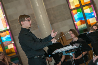 A conductor dressed in black leads a musical ensemble inside a Minnesota church with colorful stained glass windows.