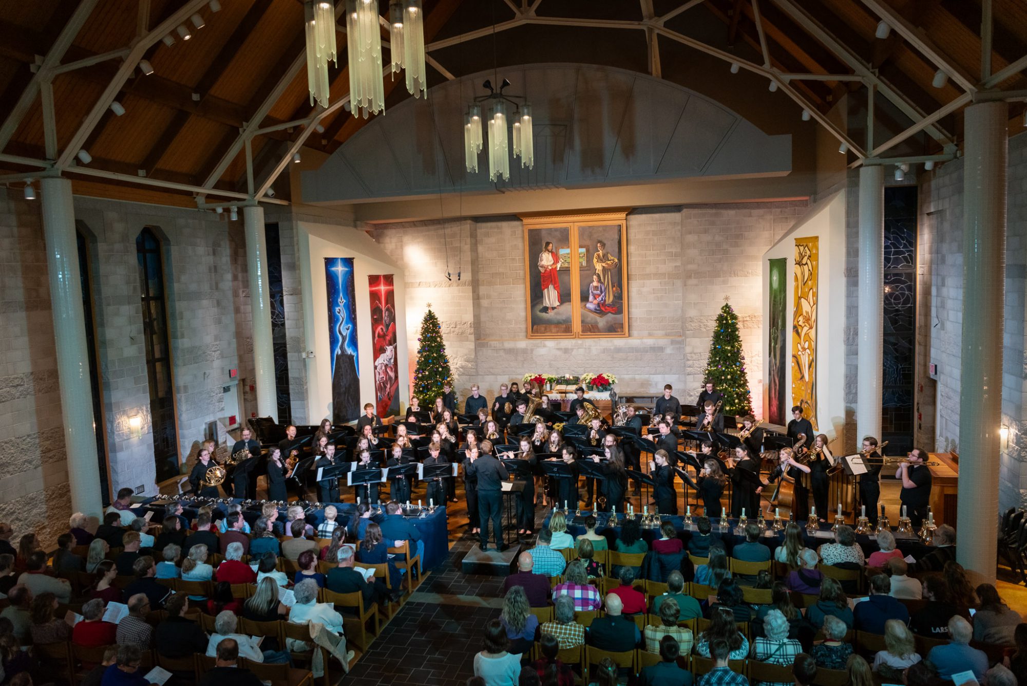A band performs in a decorated church in Minnesota, with Christmas trees in the background and an audience from the campus community.