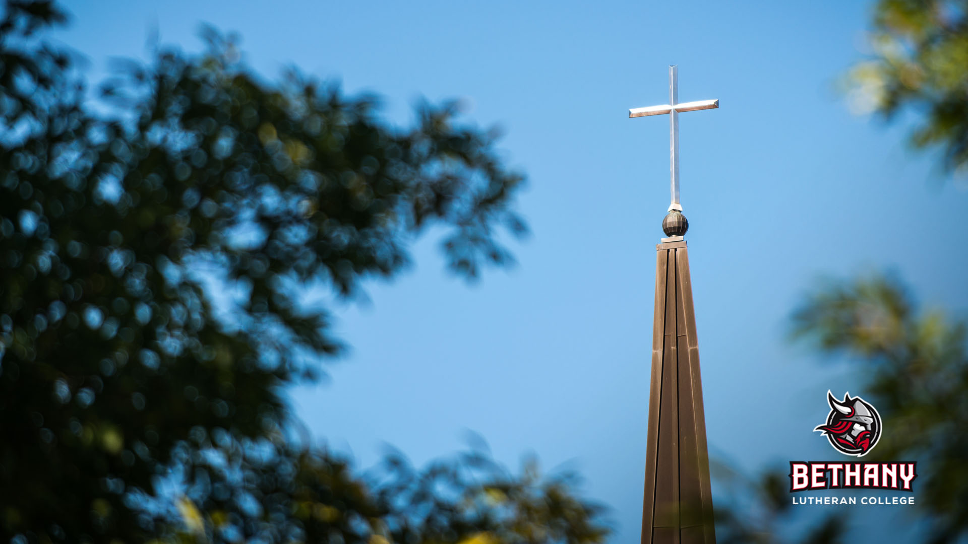 A church steeple with a cross set against a blue sky and framed by tree branches, representing the campus community at Bethany Lutheran College in Mankato.