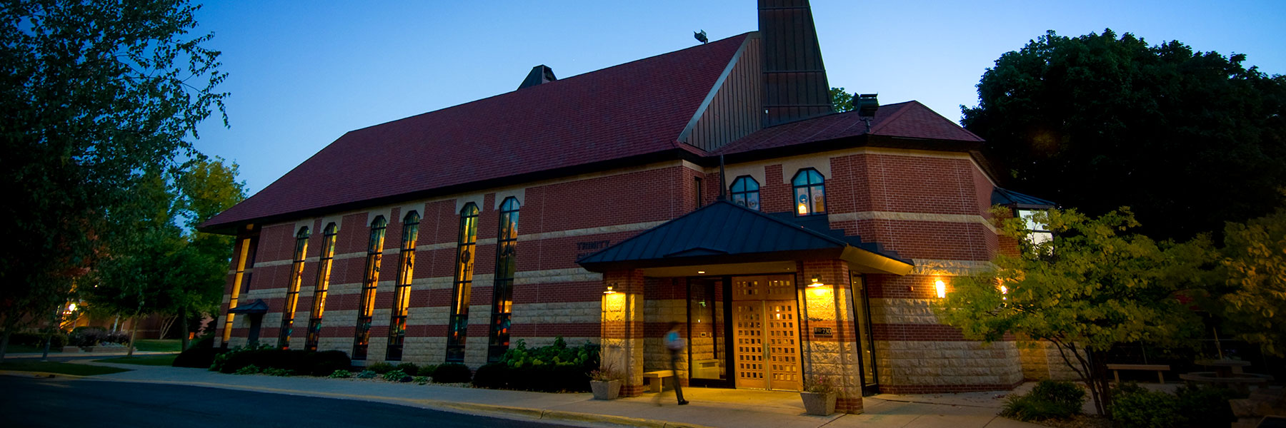 A brick building with tall windows and a peaked roof glows with exterior lights at dusk, nestled among trees on the Bethany Lutheran College campus.