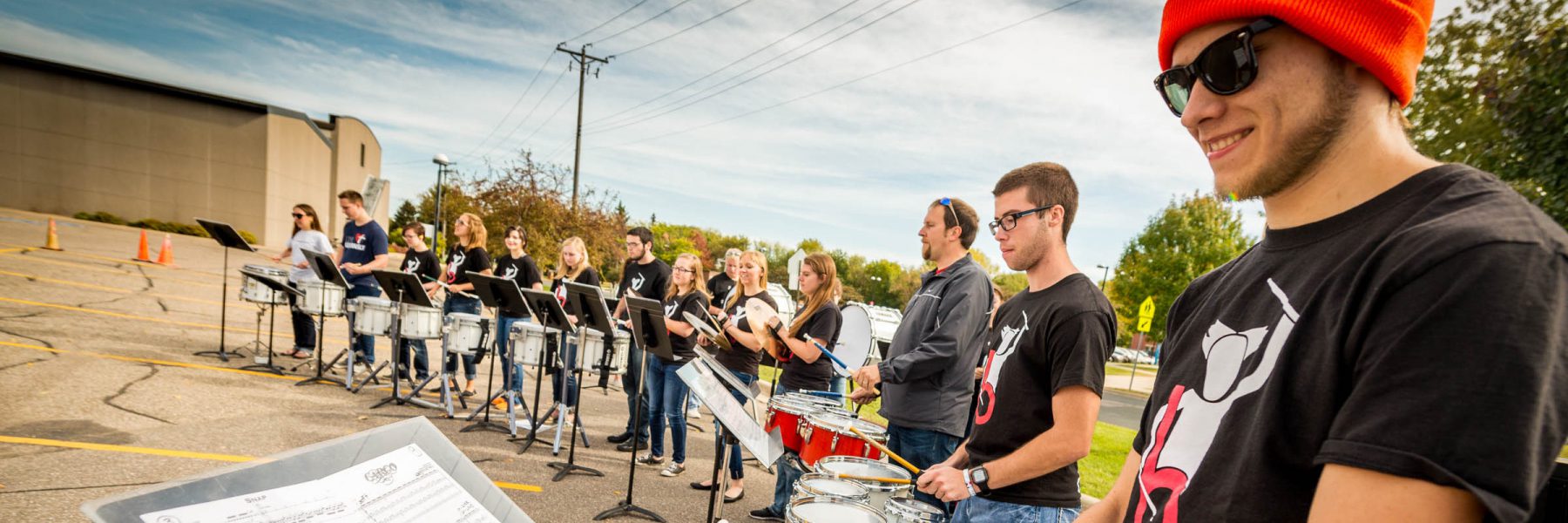 A group of students wearing black shirts play drums outdoors, fostering campus community. One student wears sunglasses and an orange beanie.