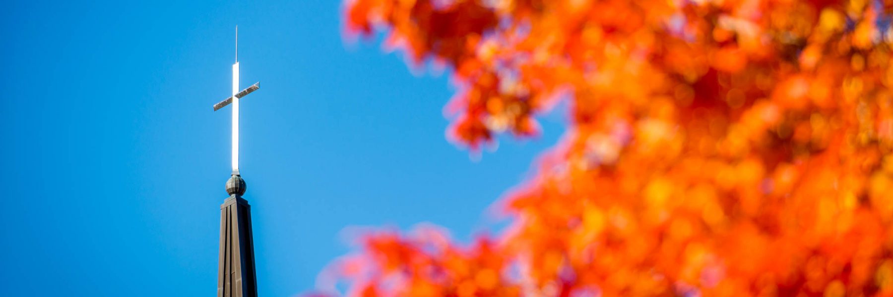 A church steeple with a cross in Mankato, Minnesota, is framed by vibrant orange autumn leaves against a clear blue sky.