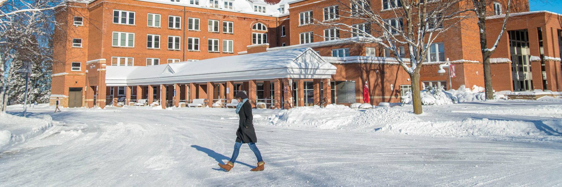 A person wearing a winter coat and boots walks across a snowy street in front of Bethany Lutheran College’s red-brick building on a sunny day.