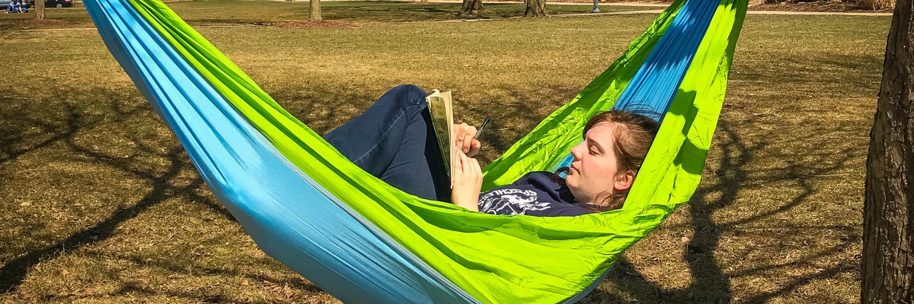 A person relaxing in a blue and green hammock outdoors on campus, reading a book and participating in student activities.