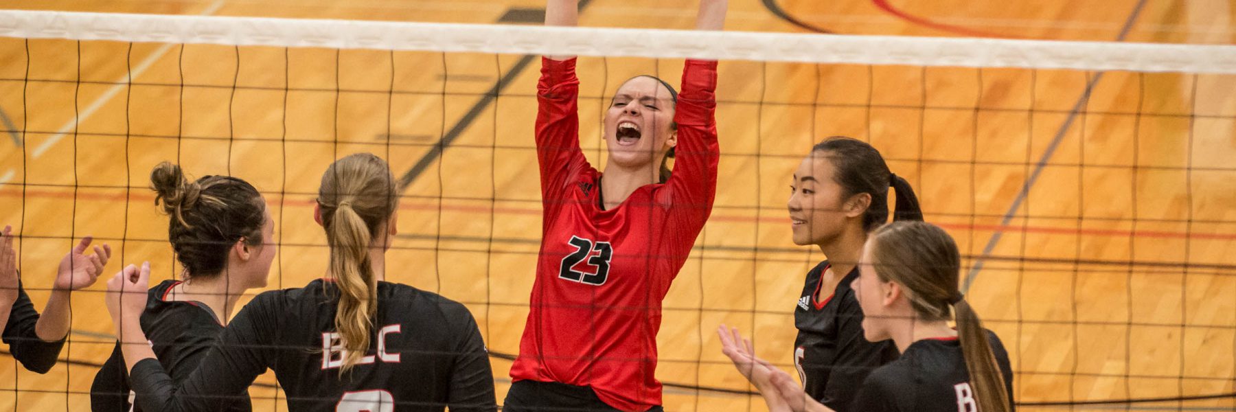 A Mankato volleyball player in a red jersey celebrates with teammates at the net on an indoor court.