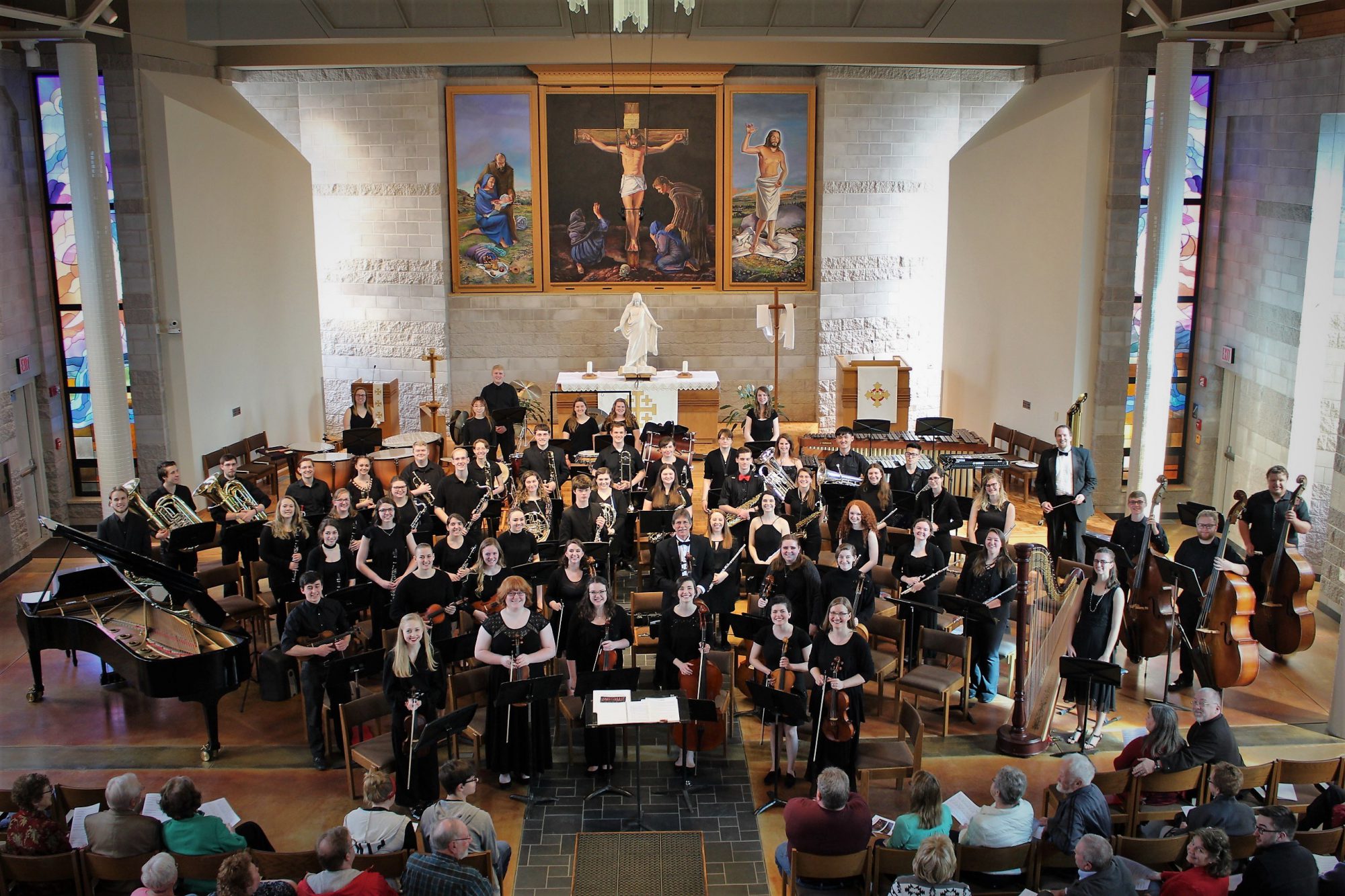 A large orchestra performs at Bethany Lutheran College in Mankato, with religious artwork displayed in the background.