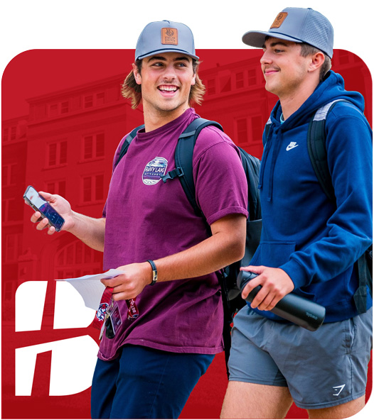 Two Bethany Lutheran College students, both wearing hats and backpacks, smile as they walk in front of a red building outline on campus.