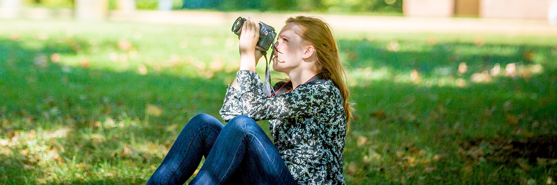 A woman sits on the grass in sunlit Mankato, Minnesota, looking upward and taking a photo with her camera.