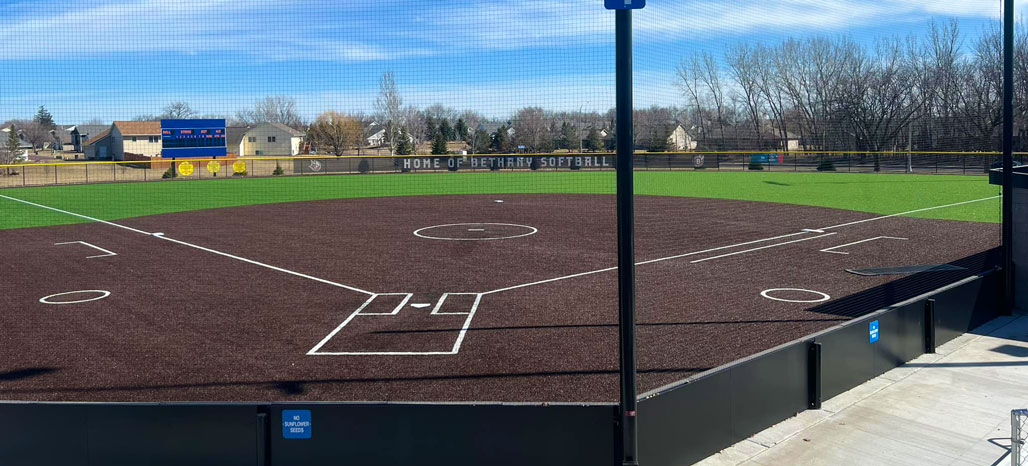 An empty softball field at Bethany Lutheran College, featuring brown turf and a fence displaying the words “Home of Bethany Softball.”.