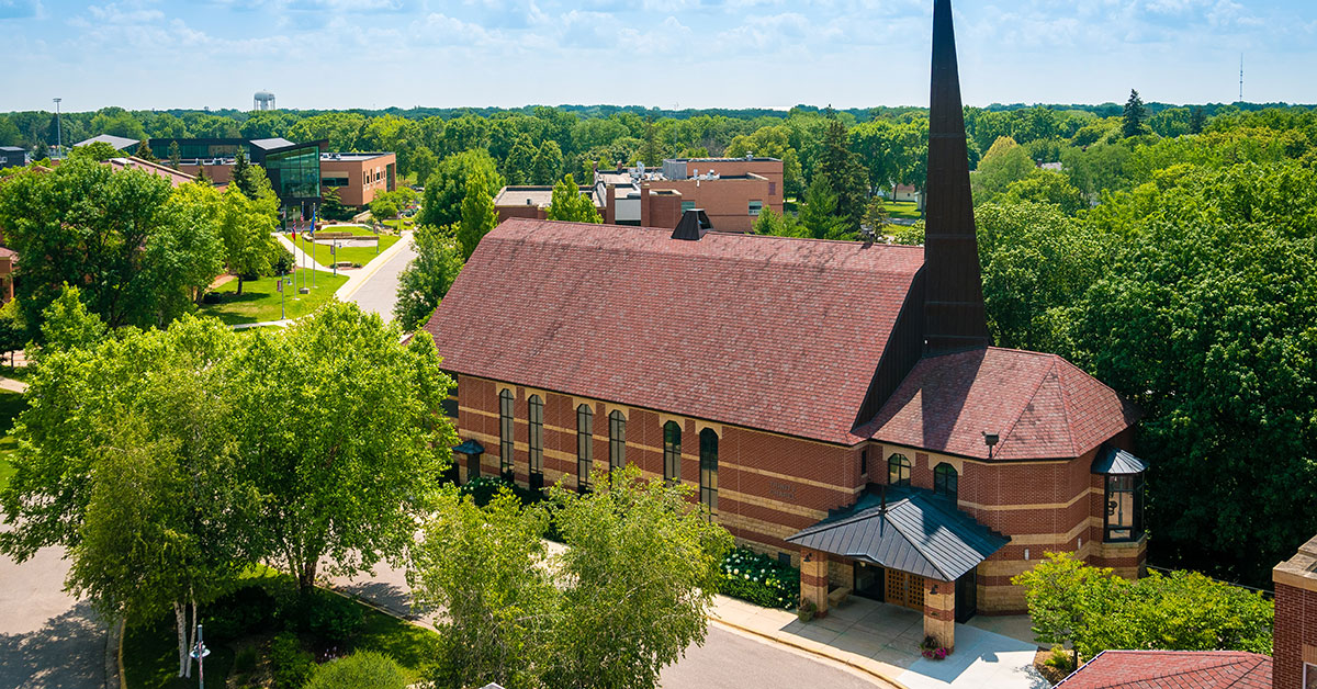 A brick chapel with a tall steeple is surrounded by green trees and campus buildings at Bethany Lutheran College on a sunny day.