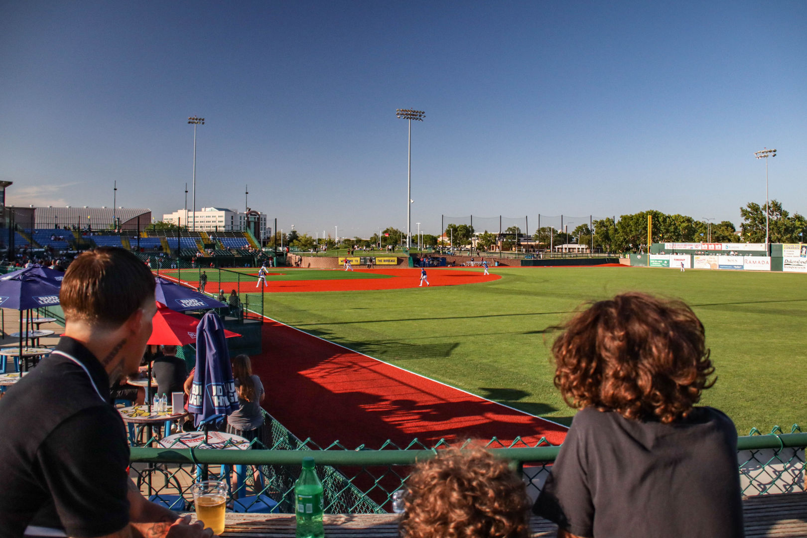 Members of the campus community watch a baseball game behind a fence on a sunny day, with a clear blue sky overhead.
