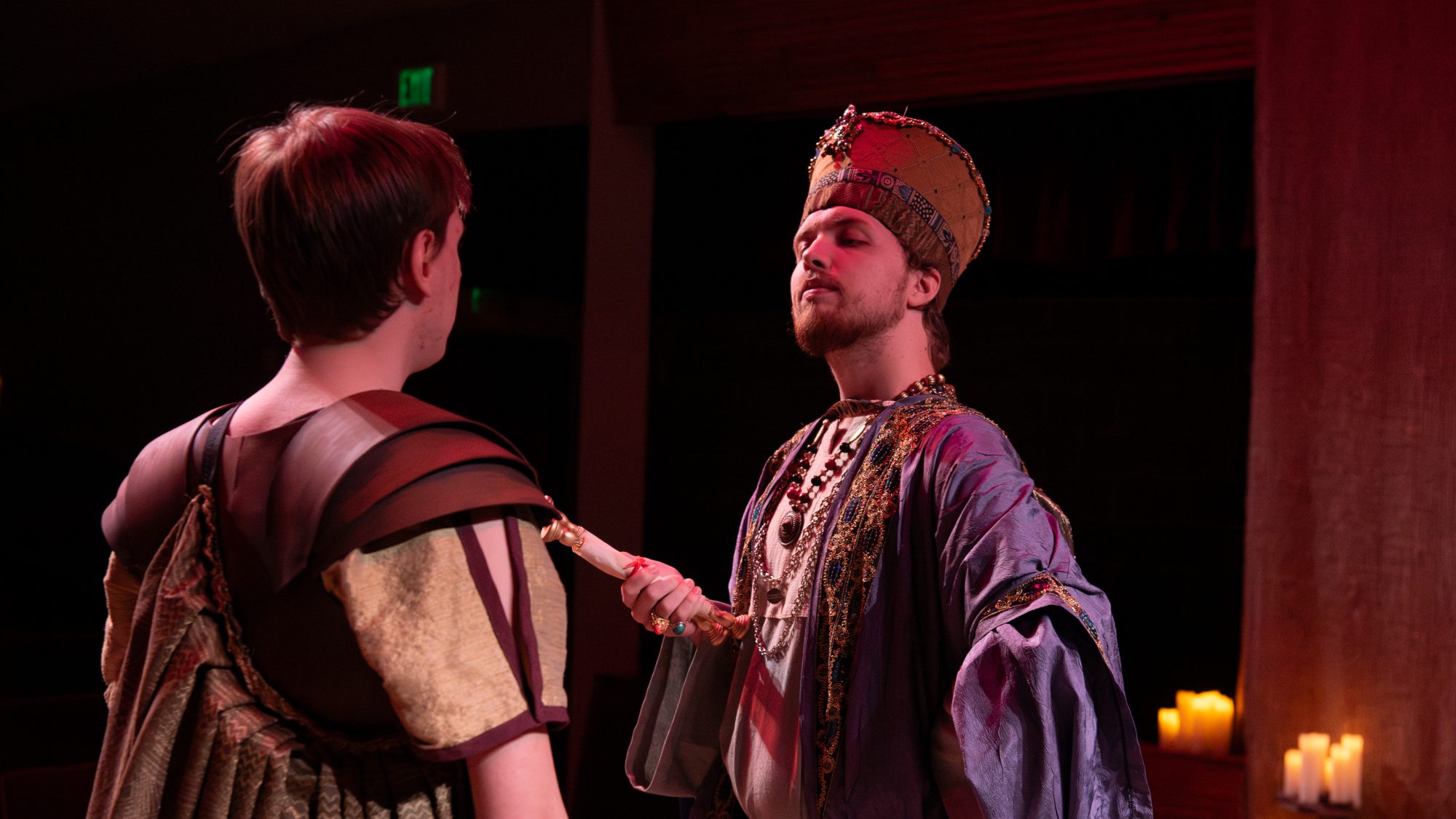 Two actors in historical costumes stand facing each other on a Minnesota stage; one, dressed in royal attire, holds a scepter to the chest of the other.