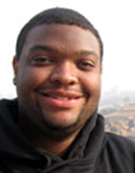 A man smiling outdoors in Mankato, Minnesota, wears a black hoodie. The background shows a blurred landscape.