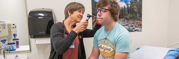 A healthcare professional examines a Minnesota student’s ear in a medical office with nature-themed wall art.