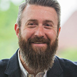 Ben Kohls, Director of Graduate Studies in Clinical Mental Health Counseling – Graduate Programs Department Online Instruction for the Academic Affairs Department, smiles in front of a blurred green background.