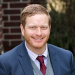 A man with short red hair and a beard, wearing a blue suit and red tie, is smiling outdoors on the Bethany Lutheran College campus in Mankato.