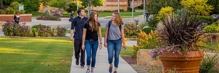 Three Bethany Lutheran College students walk together on the Mankato campus sidewalk, surrounded by vibrant green lawns, blooming flowers, and prominent campus buildings.