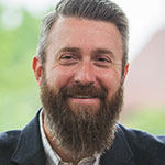A man with a thick beard and mustache, wearing a blazer and smiling, stands in front of a blurred green background at Bethany Lutheran College.