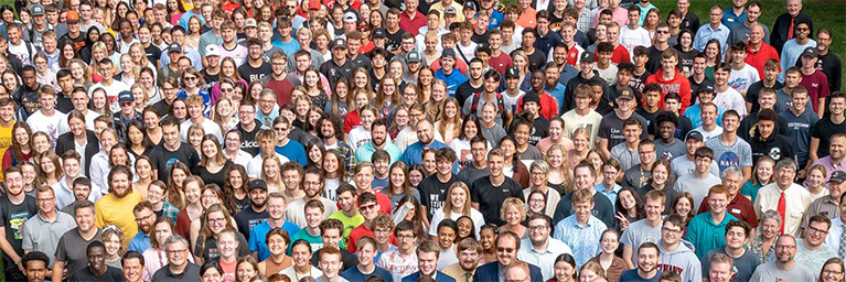 A large, diverse campus community stands closely together outdoors, smiling and facing the camera.