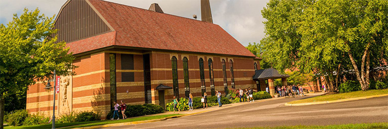 Red brick building with arched windows, surrounded by trees and members of the campus community on a sunny day in Mankato.