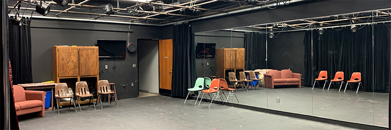 A black box theater featuring mirrors, chairs, a couch, cabinets, and overhead stage lighting.