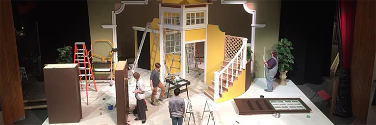 Several people assemble a colorful stage set in a theater, which features stairs, doors, and various props.