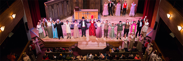 A theater cast in period costumes stands on stage, holding hands and bowing to the audience after a performance.