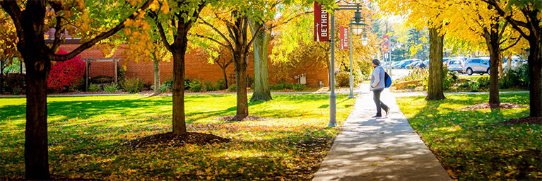 A person walks on a sunlit Mankato campus, surrounded by green grass, autumn leaves, and members of the campus community.