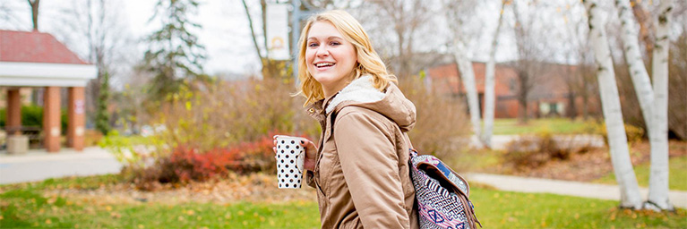 A smiling young woman with a backpack and coffee cup walks outside on the Bethany Lutheran College campus.